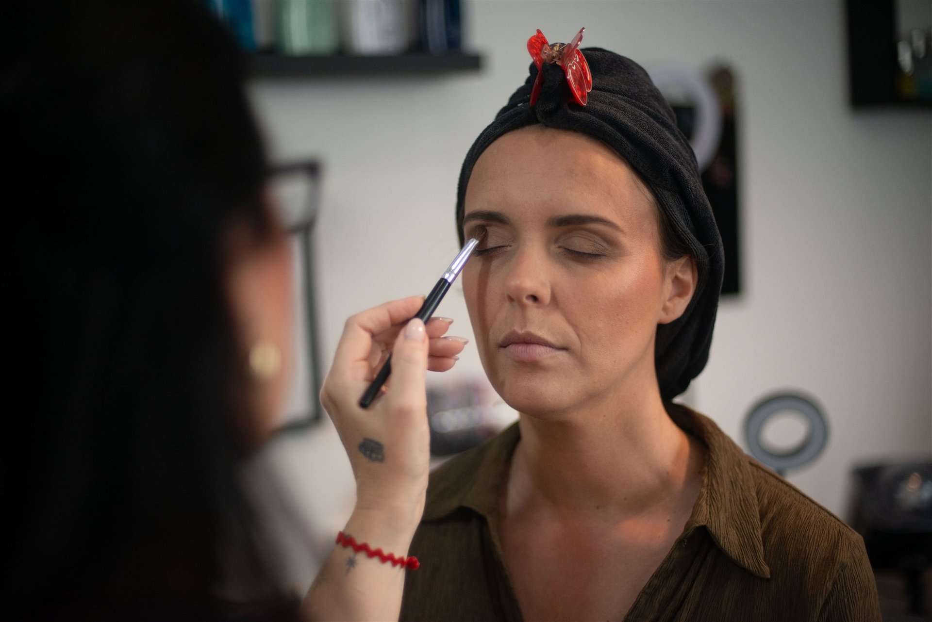 Magali applique l'eye-liner sur la paupière d'Amandine — profil, tête dans serviette noire, pince cerise rouge