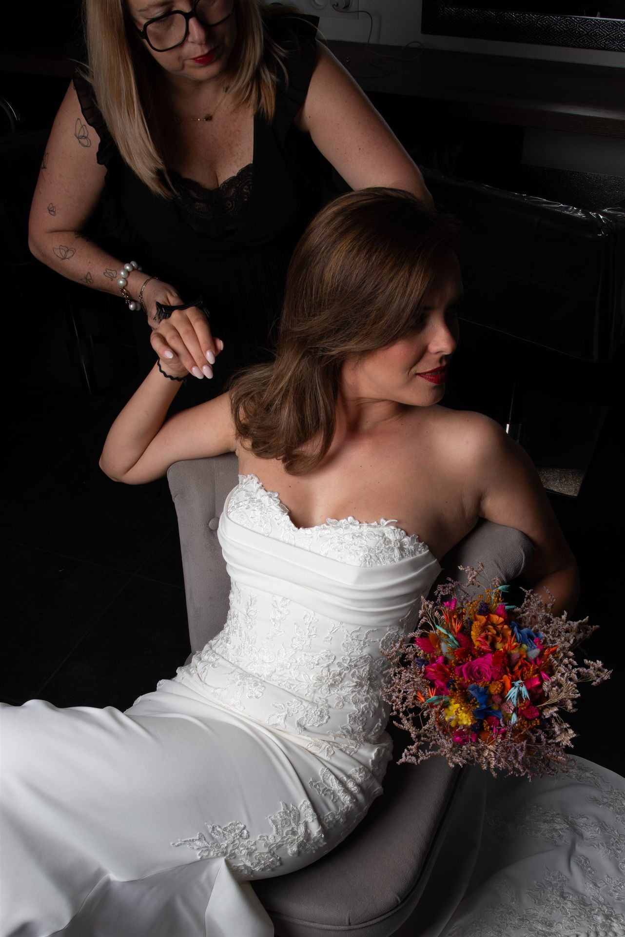 Annabel travaille la coiffure de la mariée dans un fauteuil, bouquet coloré fuchsia et orange — shooting mariage Perpignan